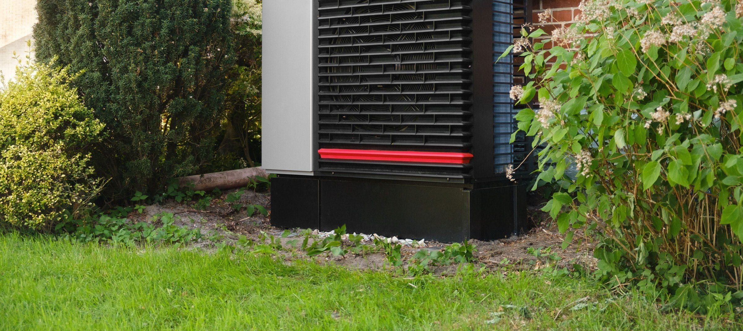 An air source heat pump beside a brick house, planting around it