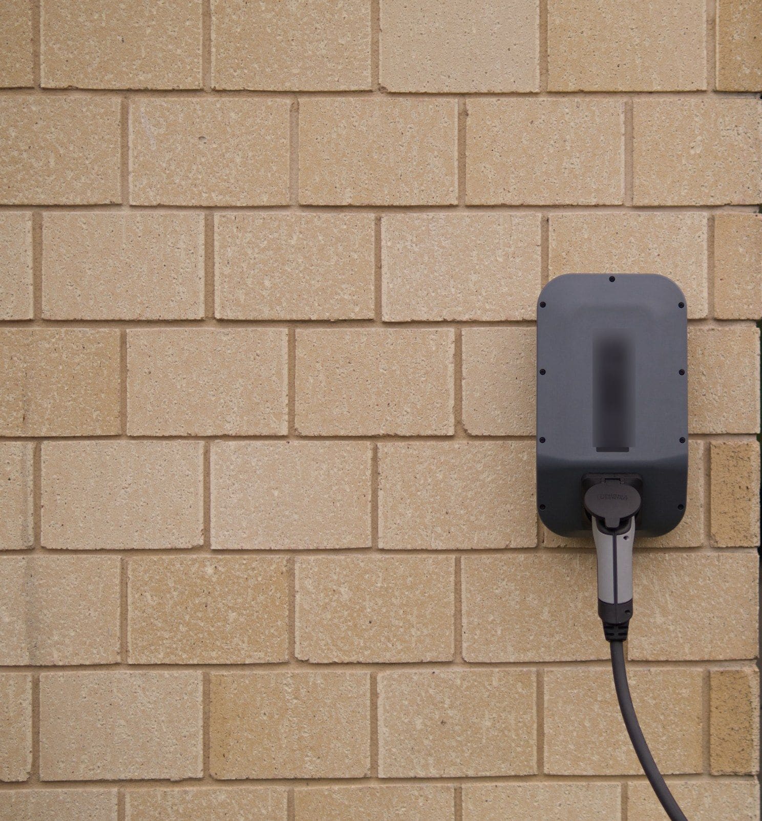 A wall-mounted electric car charger on a buff brick wall, its cable plugged in
