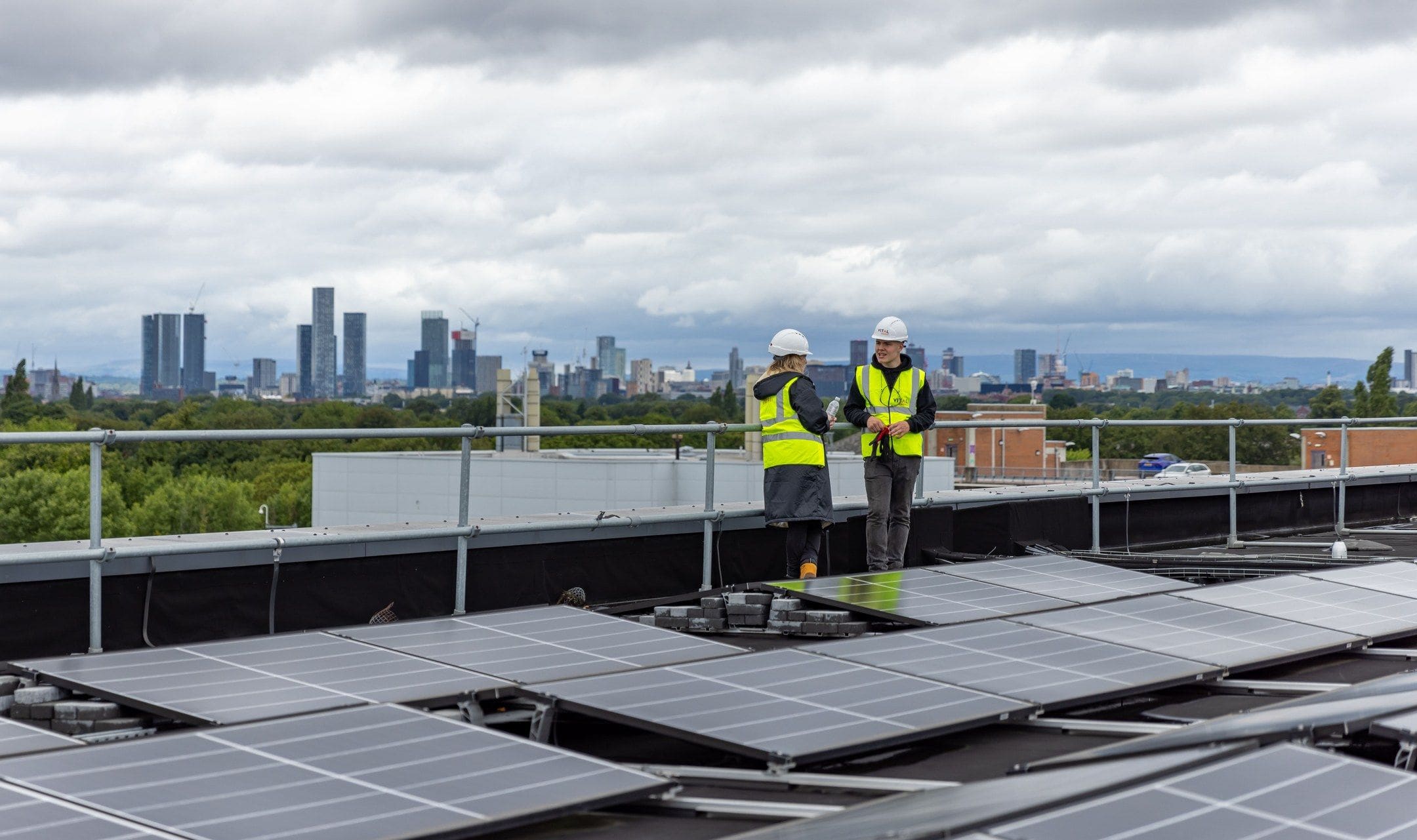 Two installers in hard hats checking solar panels on a flat roof, a city skyline beyond